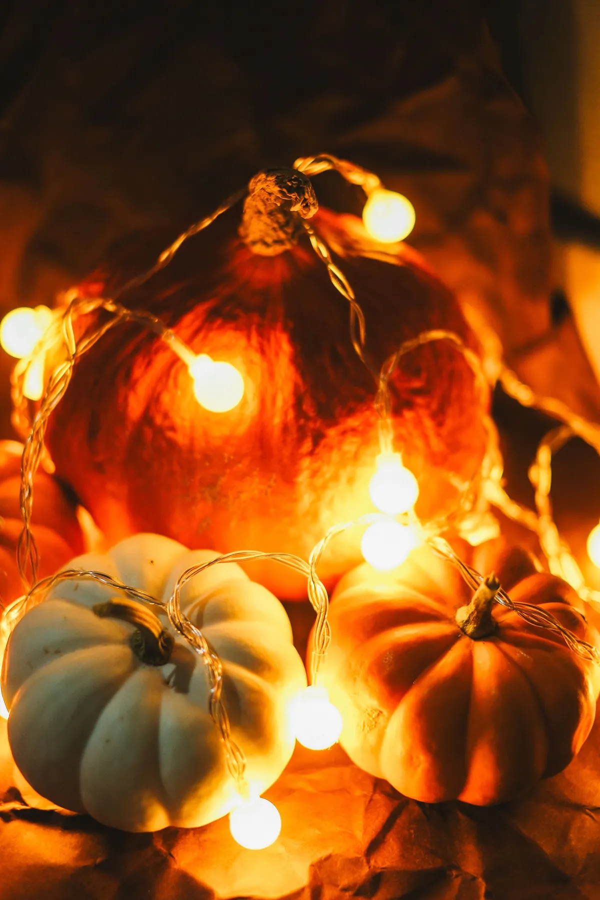 Orange and pale green pumpkins wrapped in glowing warm-white fairy lights against a dark background