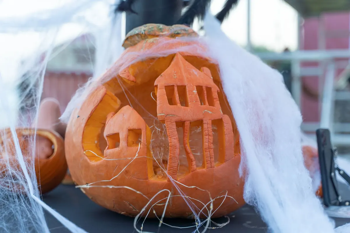 Large pumpkin carved in relief with a tall haunted house and a smaller house, draped with white cobweb