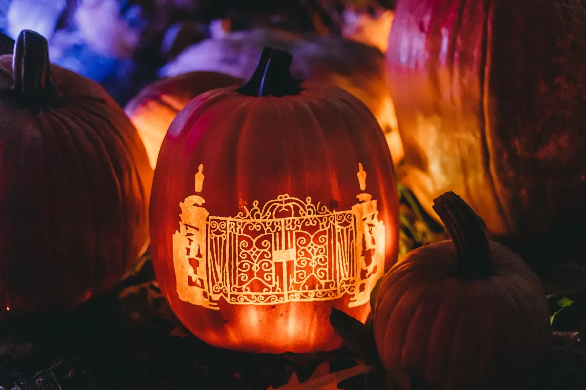 Tall glowing pumpkin carved with an ornate wrought-iron gate between two pillars, other pumpkins blurred behind