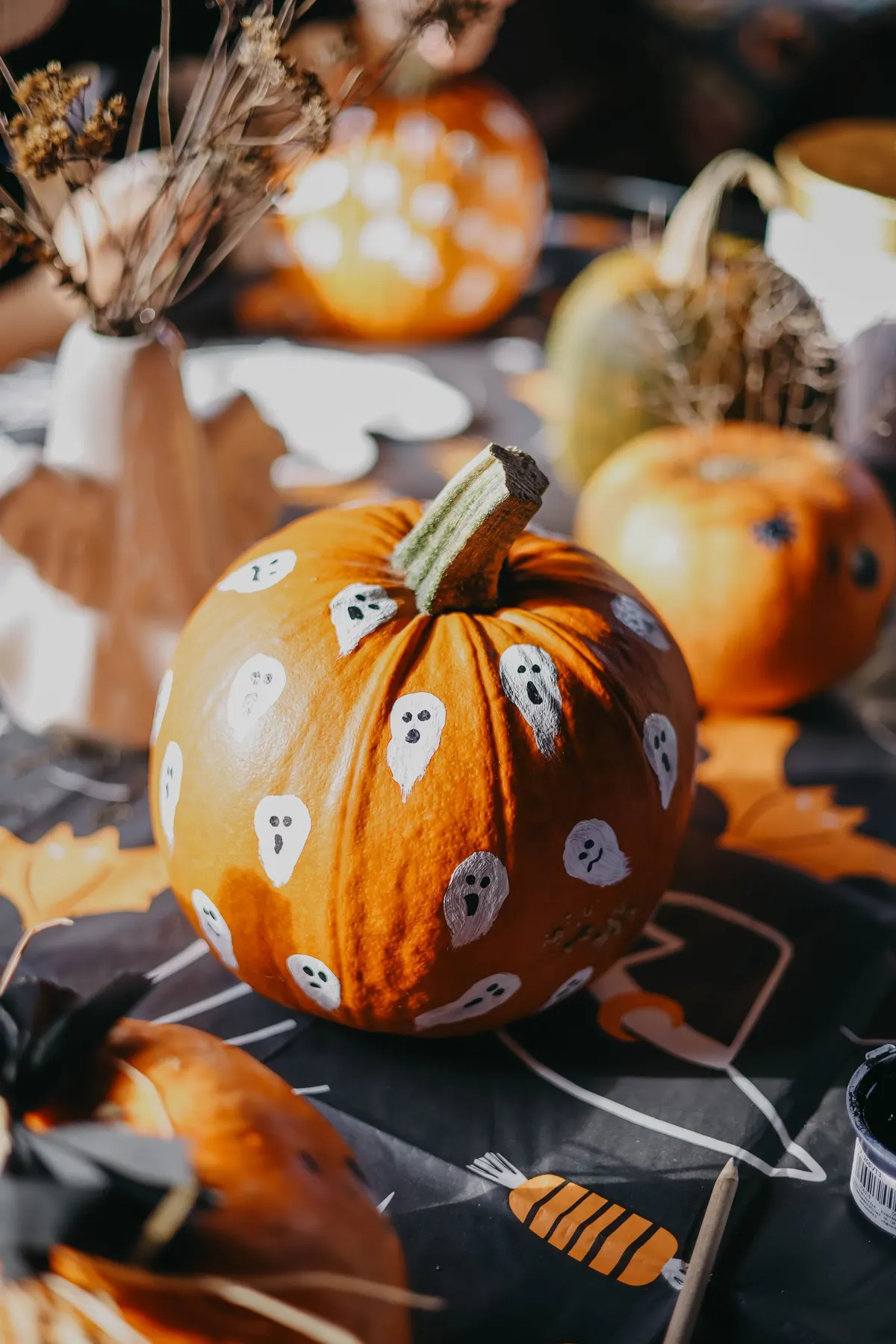 Orange pumpkin painted all over with small white ghosts with tiny dark faces, on a patterned tablecloth
