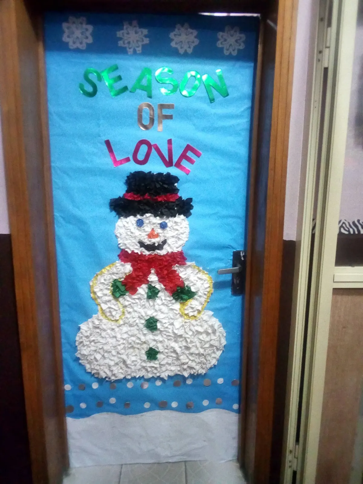 Door covered in light blue paper and snowflakes with a crumpled-paper snowman wearing a black top hat and red bow
