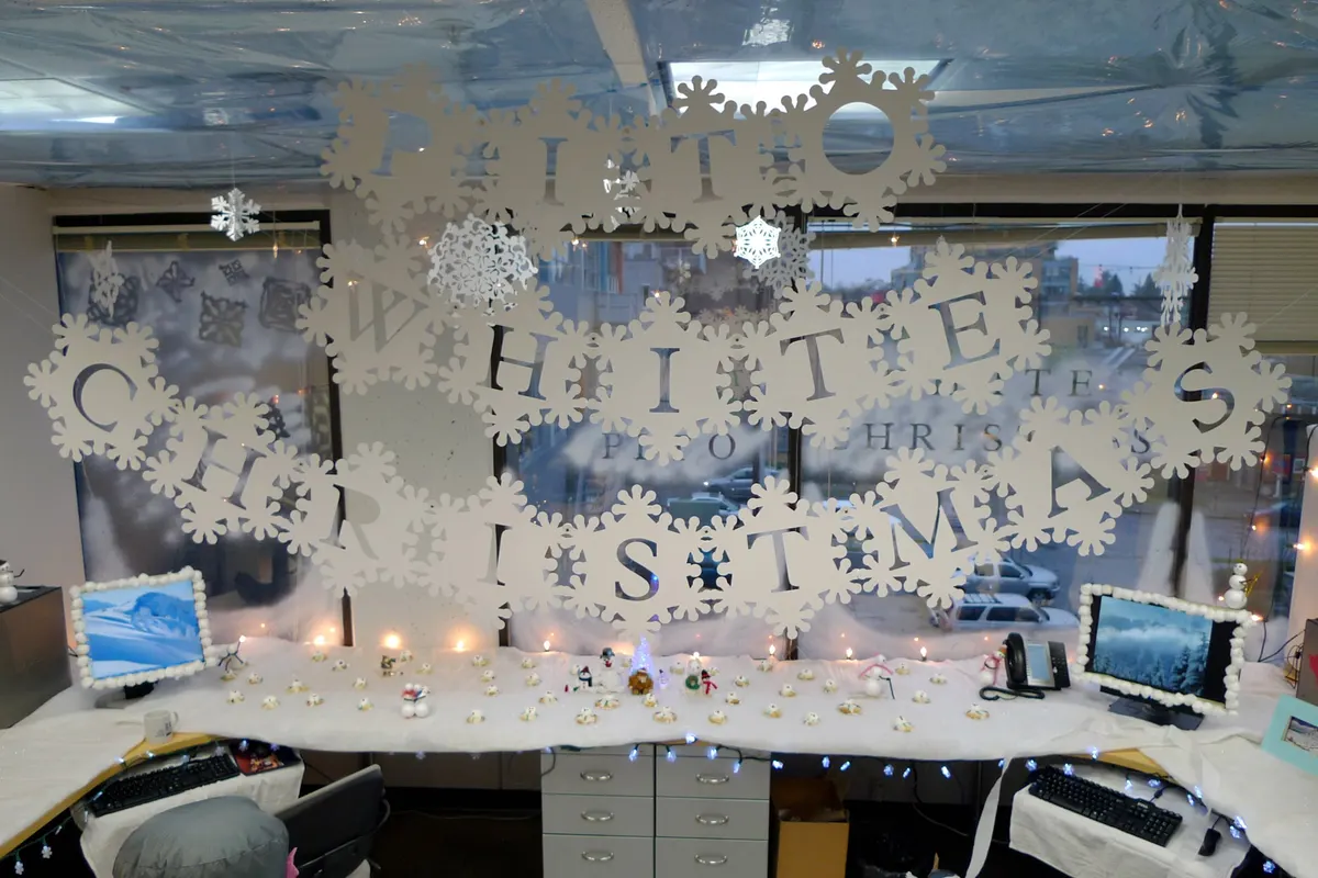 Office cubicle decorated in white with large paper snowflakes on the windows, snow-covered desks and string lights