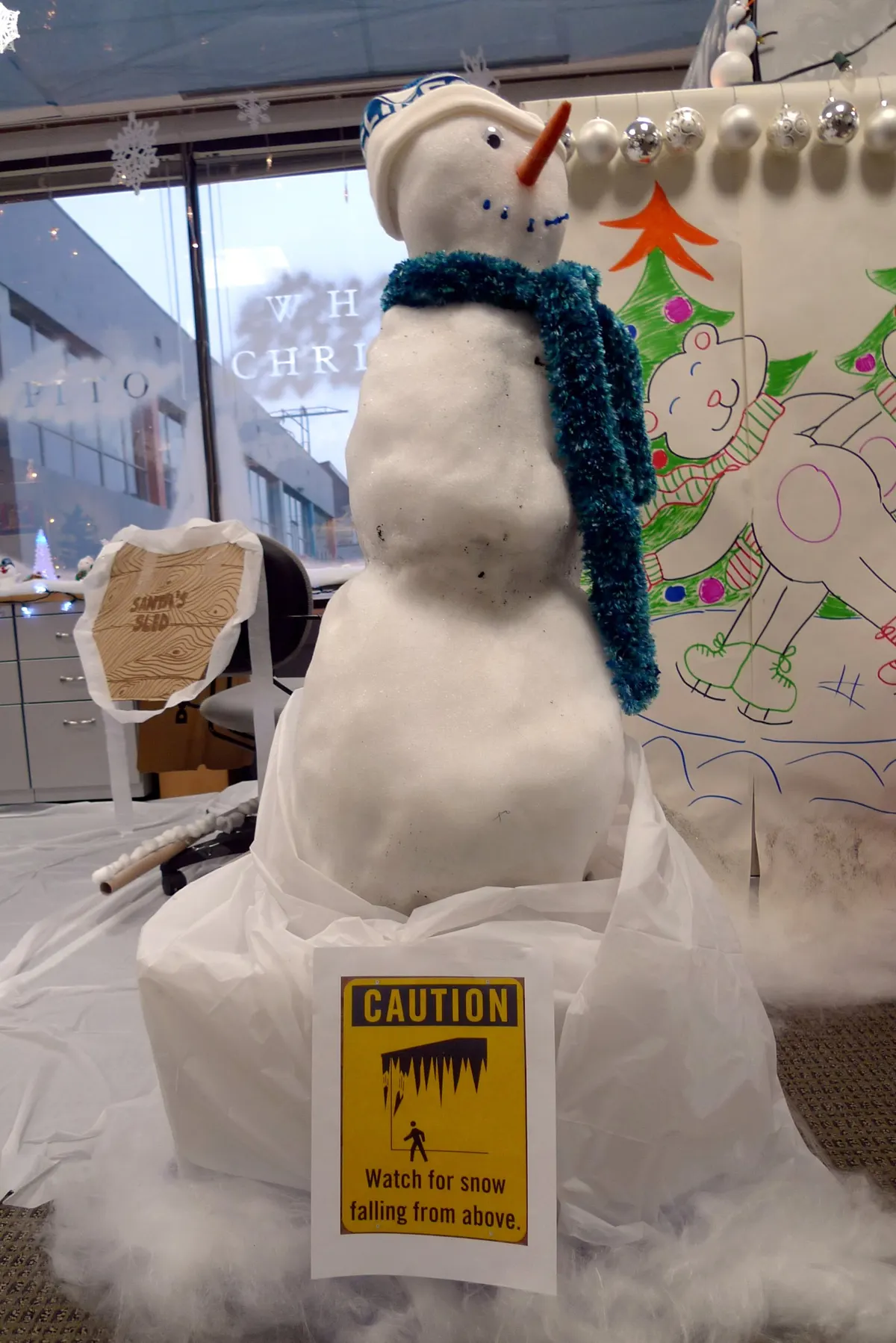 Small snowman with a teal tinsel scarf and carrot nose in front of a hand-drawn Christmas mural in an office