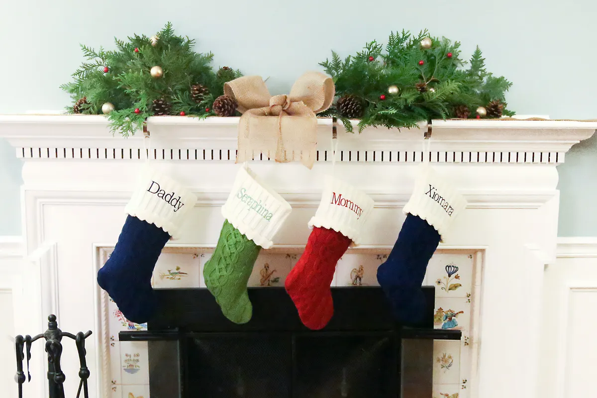 White mantel with an evergreen garland, pinecones, gold ornaments, a burlap bow and four knit stockings hanging below