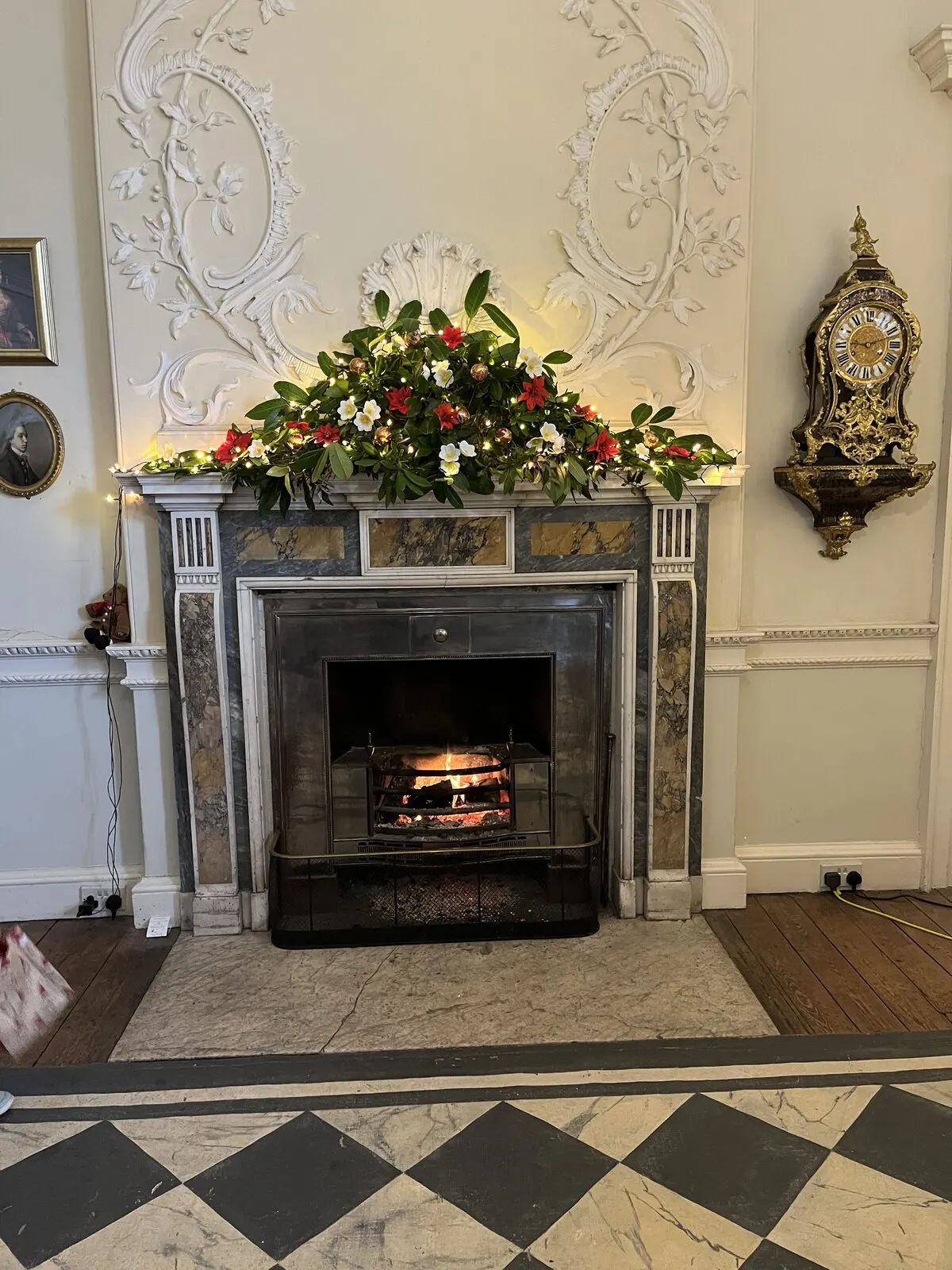 Marble mantelpiece topped with an evergreen garland, red flowers, gold baubles and warm white fairy lights