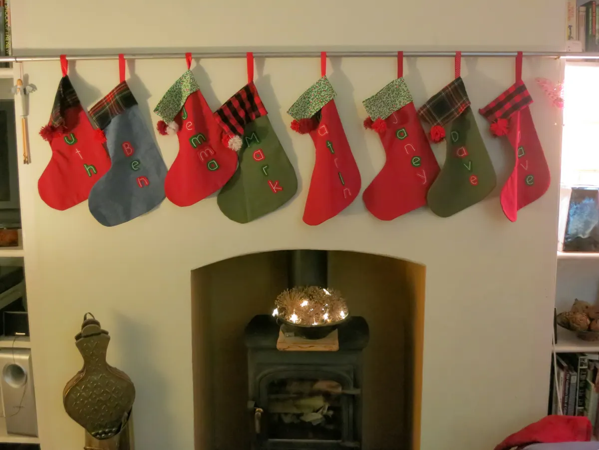 Eight handmade stockings hanging from a slim metal rail on a white chimney breast above a small wood stove