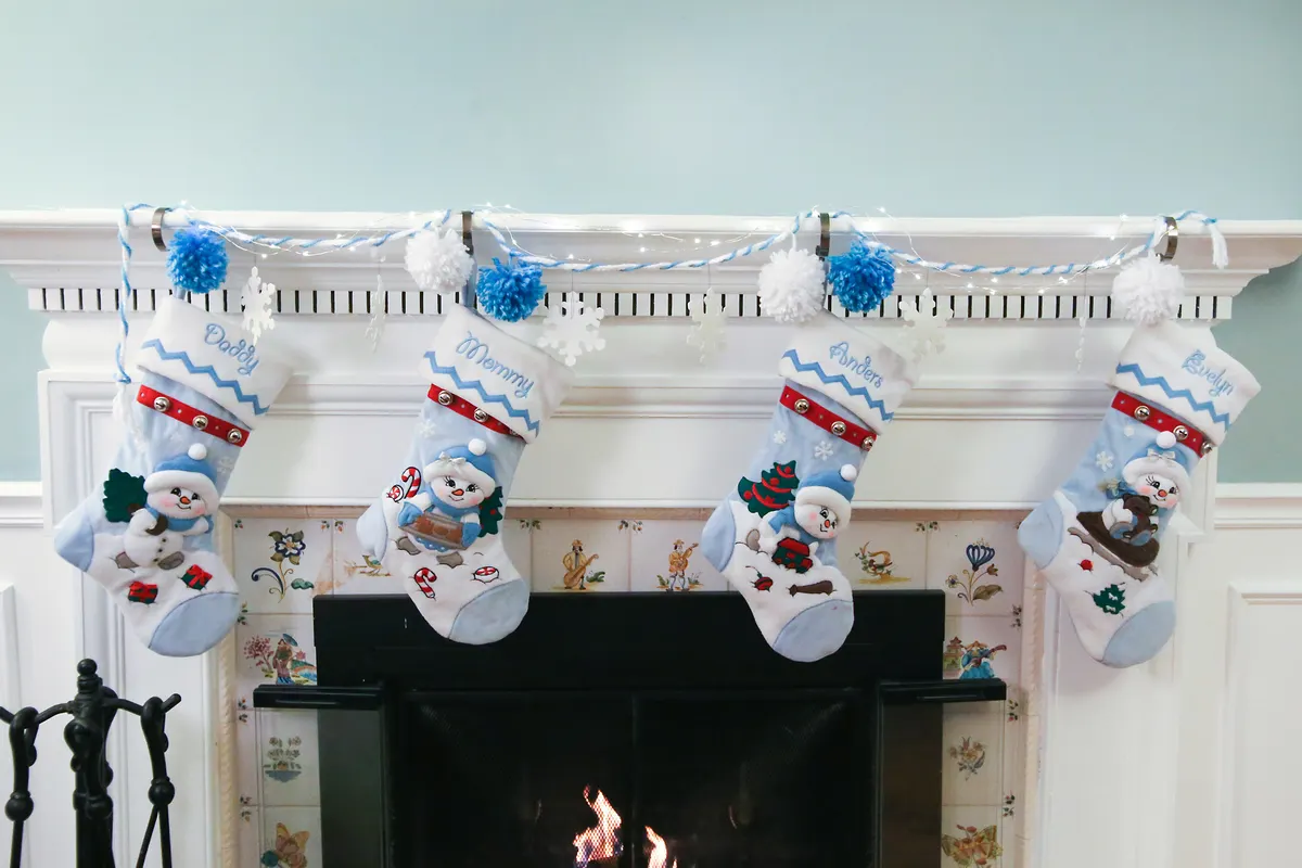 White mantel with a blue and white garland, fairy lights, pom-poms and four snowman stockings above a lit fireplace