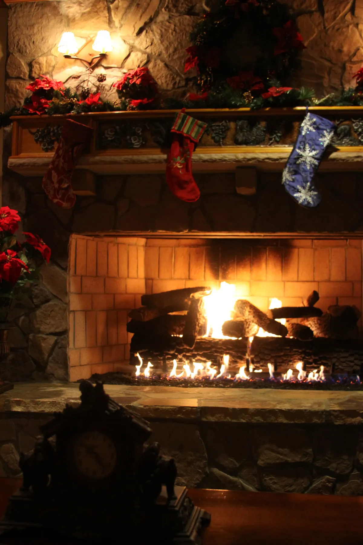 Stone fireplace with a wood mantel, evergreen and red poinsettia garland, a large wreath and three stockings above a fire