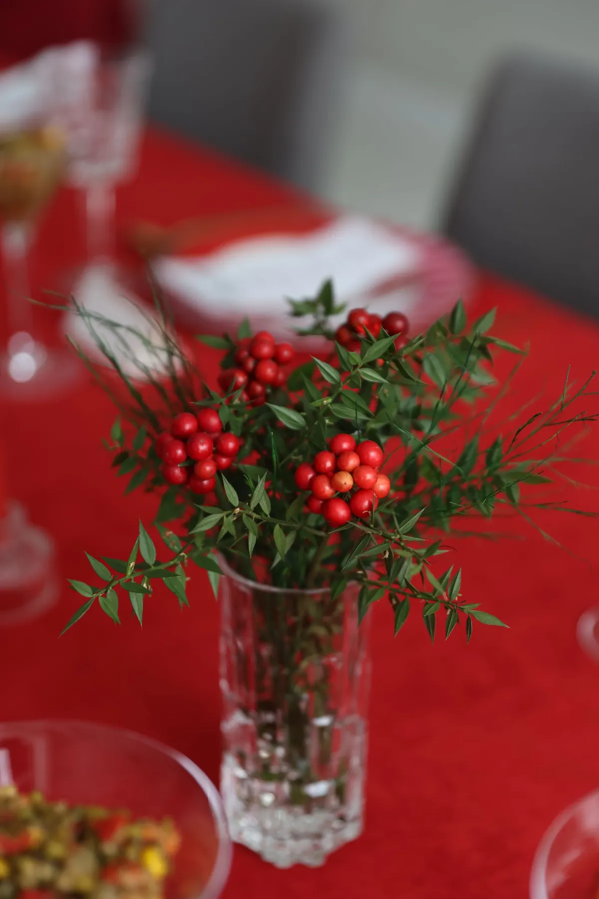 Tall cut-glass vase of green branches and red berries on a red tablecloth with clear dishes and stemware