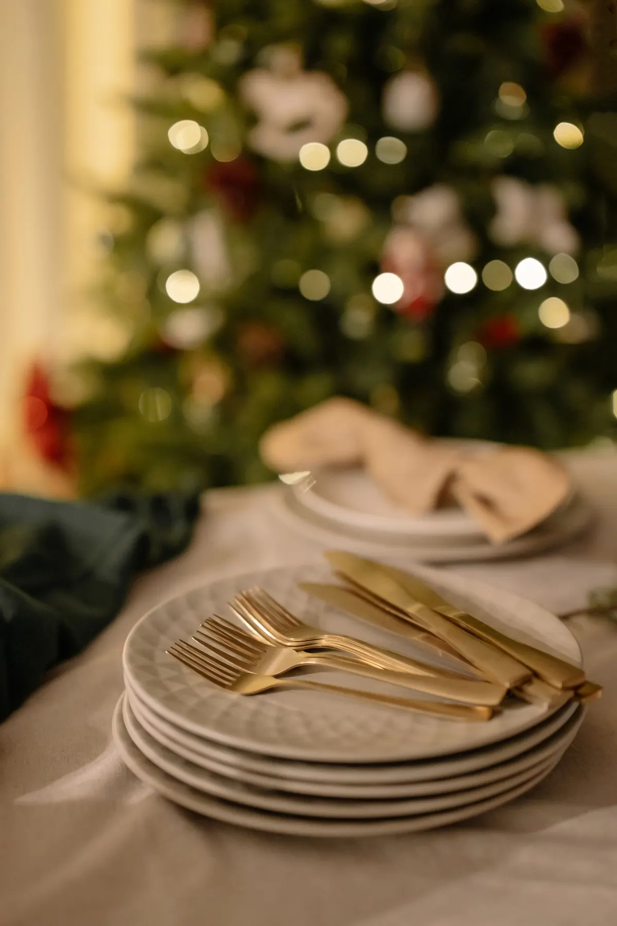 Stack of white plates holding gold forks and knives on a cream cloth with a lit Christmas tree behind