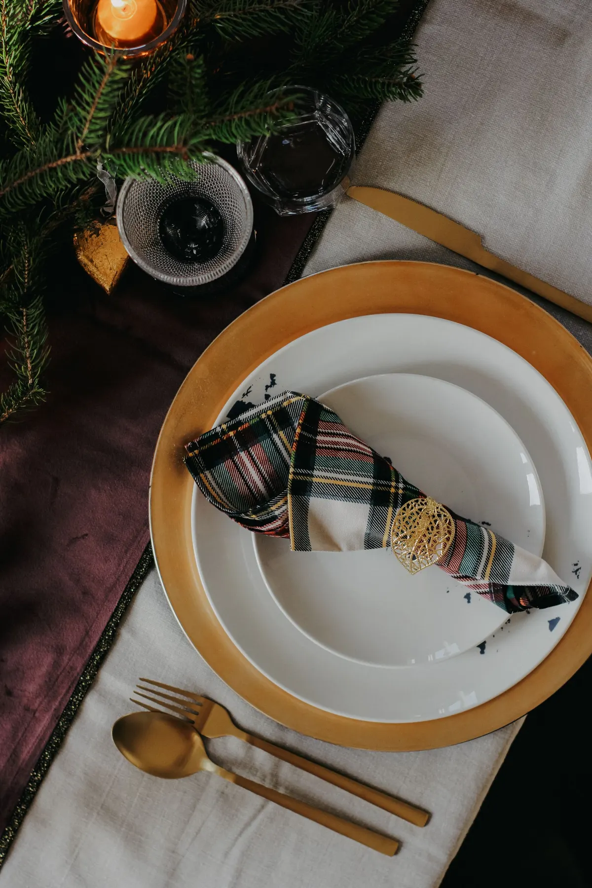 Burgundy runner on an off-white cloth with a gold charger, white plates and a tartan napkin in a gold ring