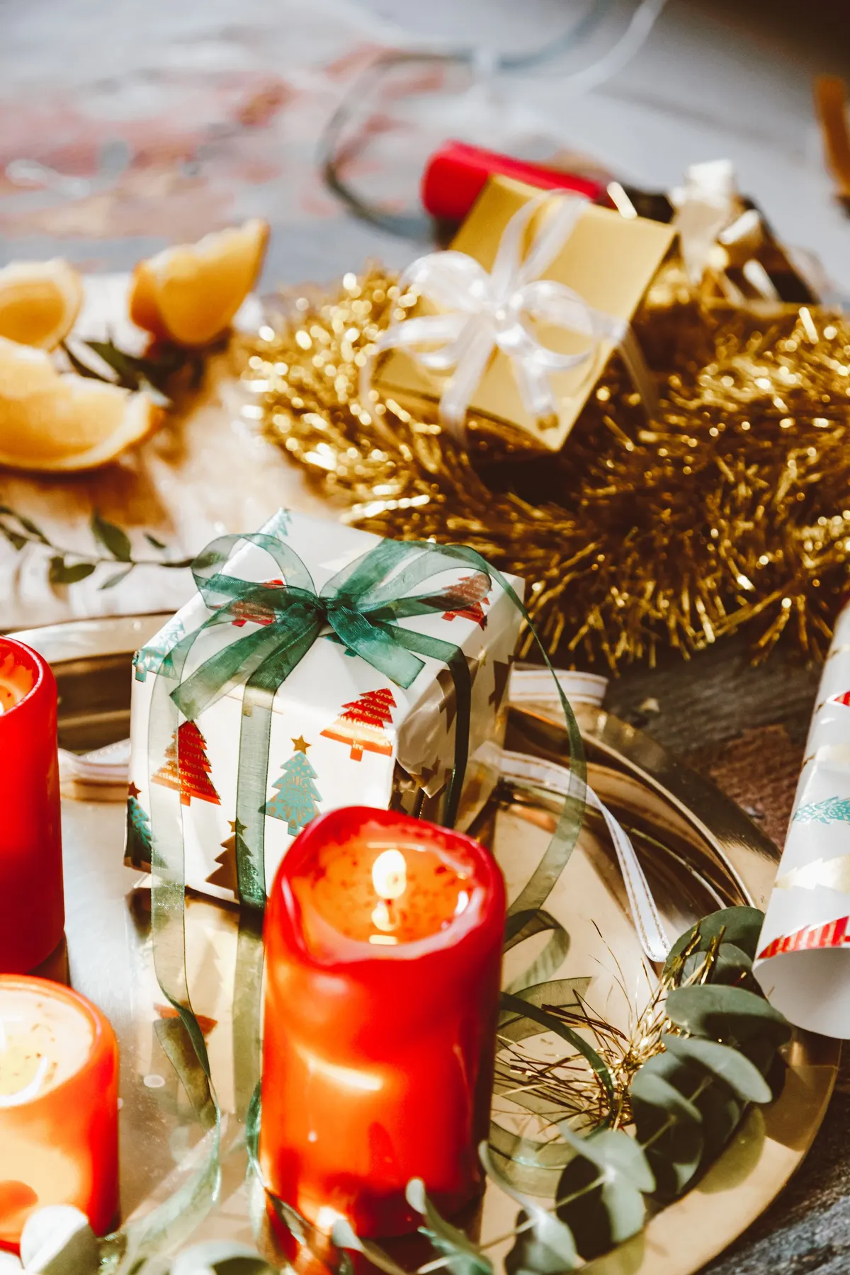 Three red pillar candles on gold trays with eucalyptus, citrus wedges, gold tinsel and two wrapped gifts