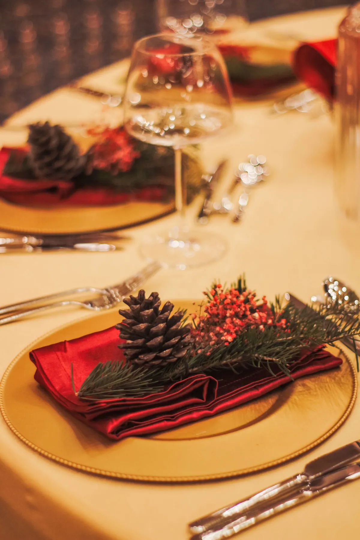 Cream tablecloth with gold chargers, red satin napkins, pinecones and evergreen sprigs beside clear wine glasses