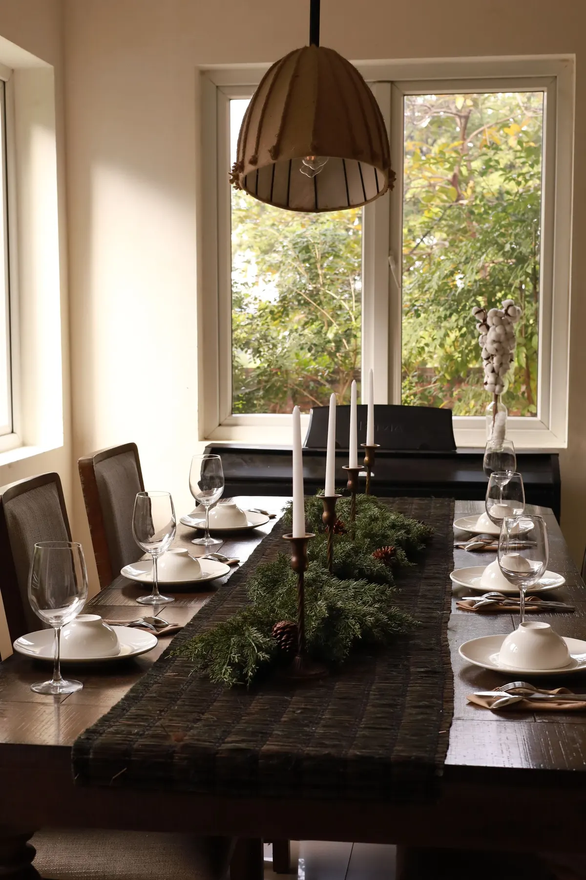 Long wooden table with a dark striped runner, six white place settings, four white tapers and evergreen