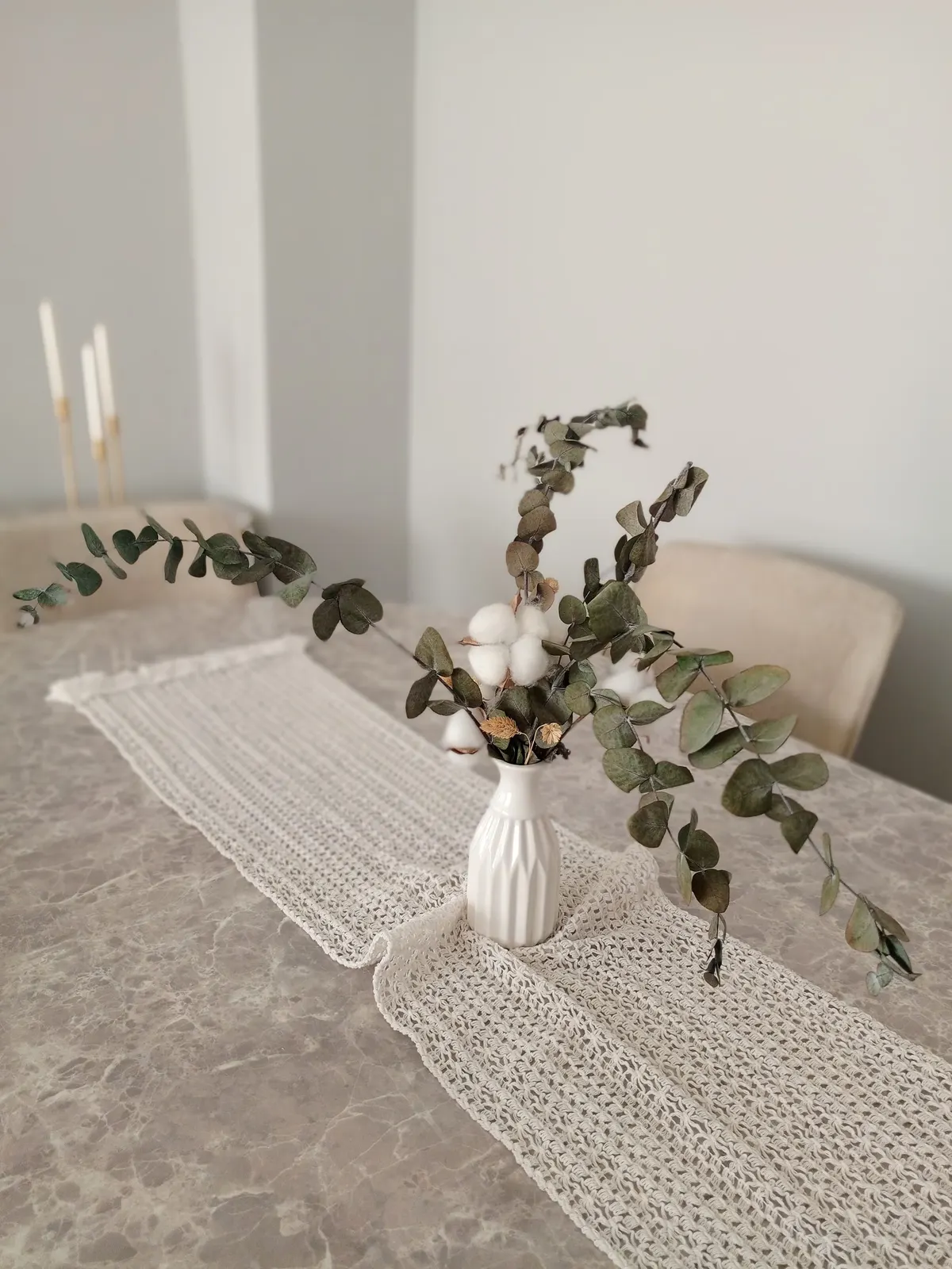 White ribbed vase of eucalyptus and cotton stems on a crocheted runner with three white tapers behind