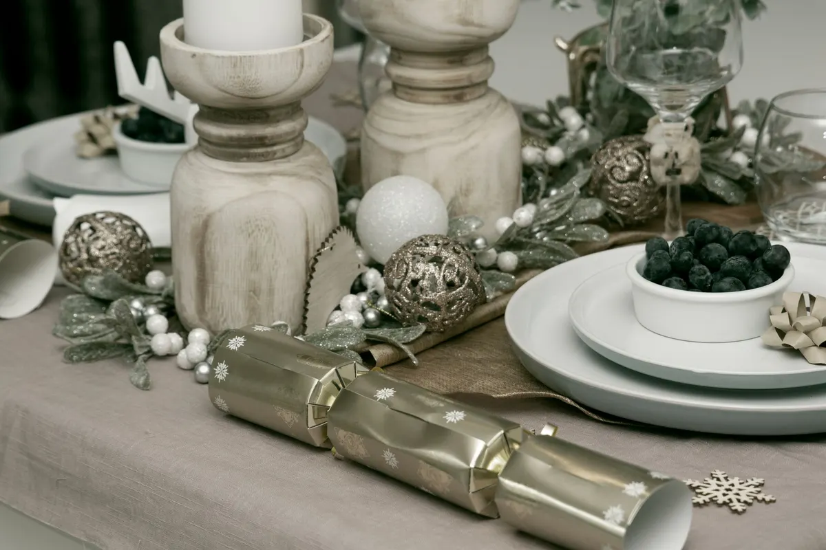 Taupe tablecloth with a gold runner, glittered greenery, silver filigree balls and wooden candleholders between two place settings