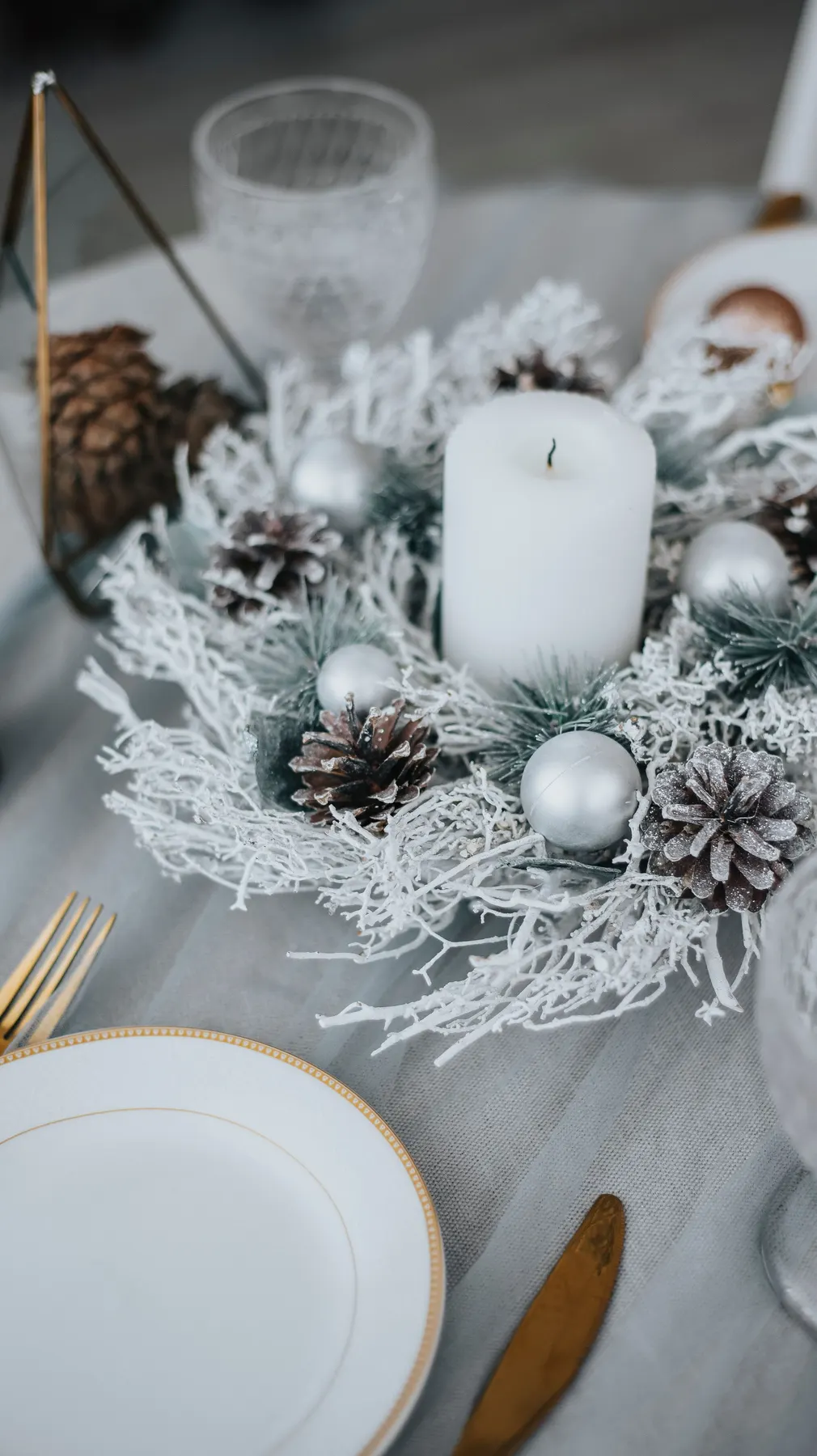 White pillar candle set in frosted evergreen branches with silver baubles and pinecones on a gray tablecloth