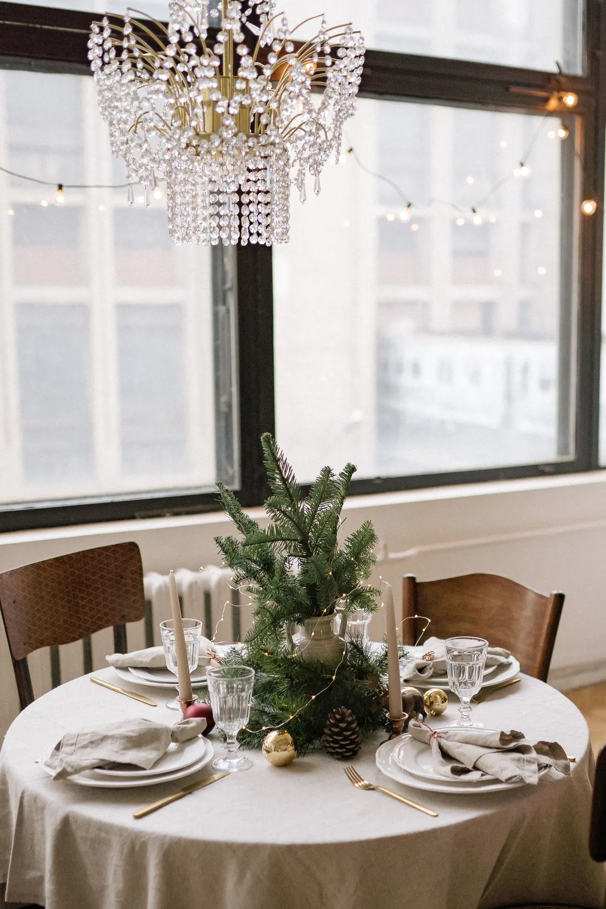 Round table with four place settings, an evergreen pitcher centerpiece, two beige tapers in copper holders and baubles
