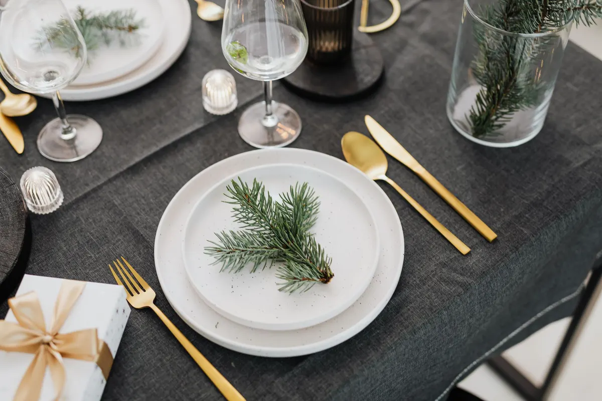 Charcoal tablecloth with stacked white plates topped by evergreen sprigs, gold cutlery and a glass vase with a branch