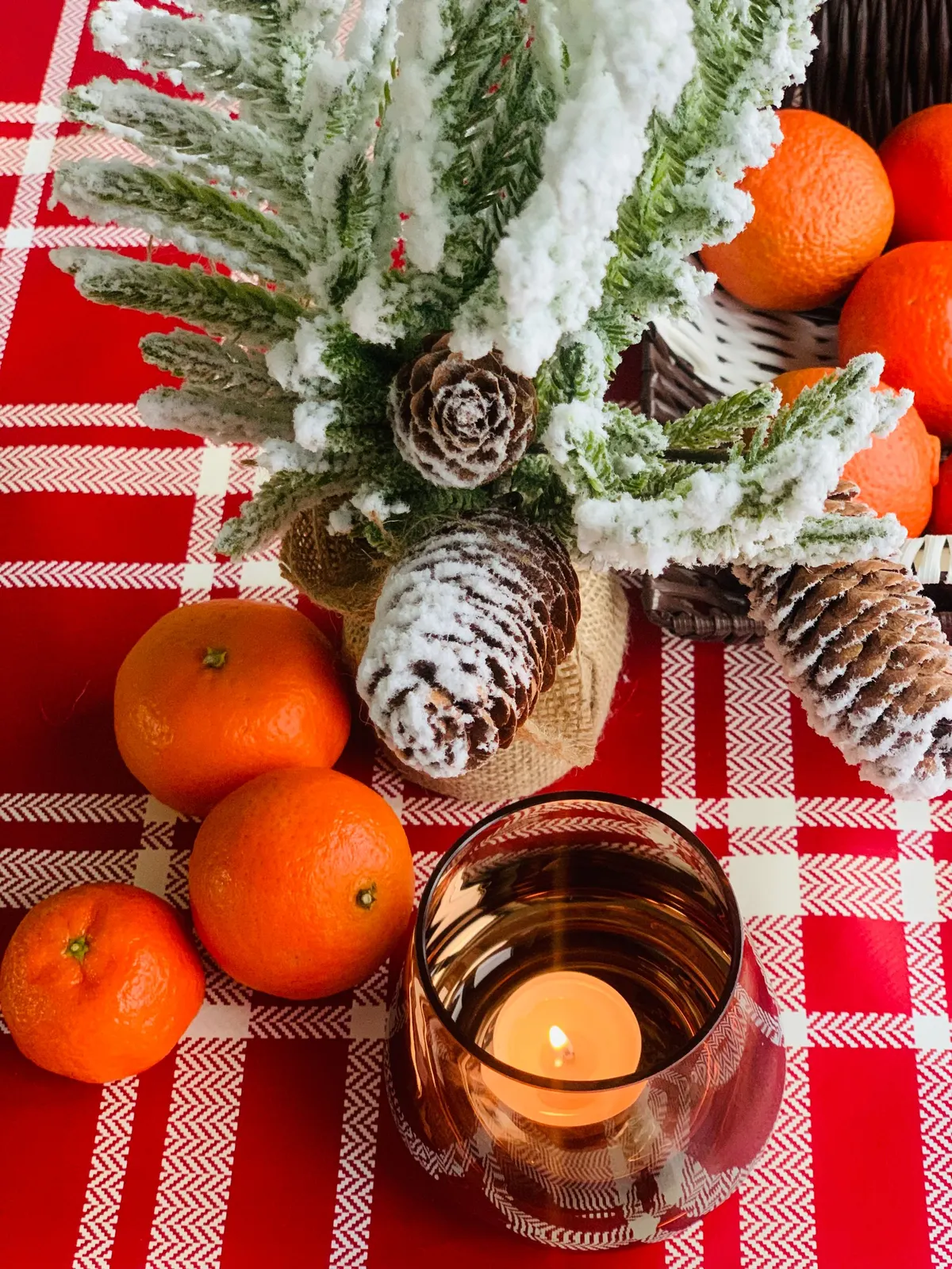 Snow-flocked evergreen in burlap with pinecones, loose oranges and a candle in amber glass on red plaid