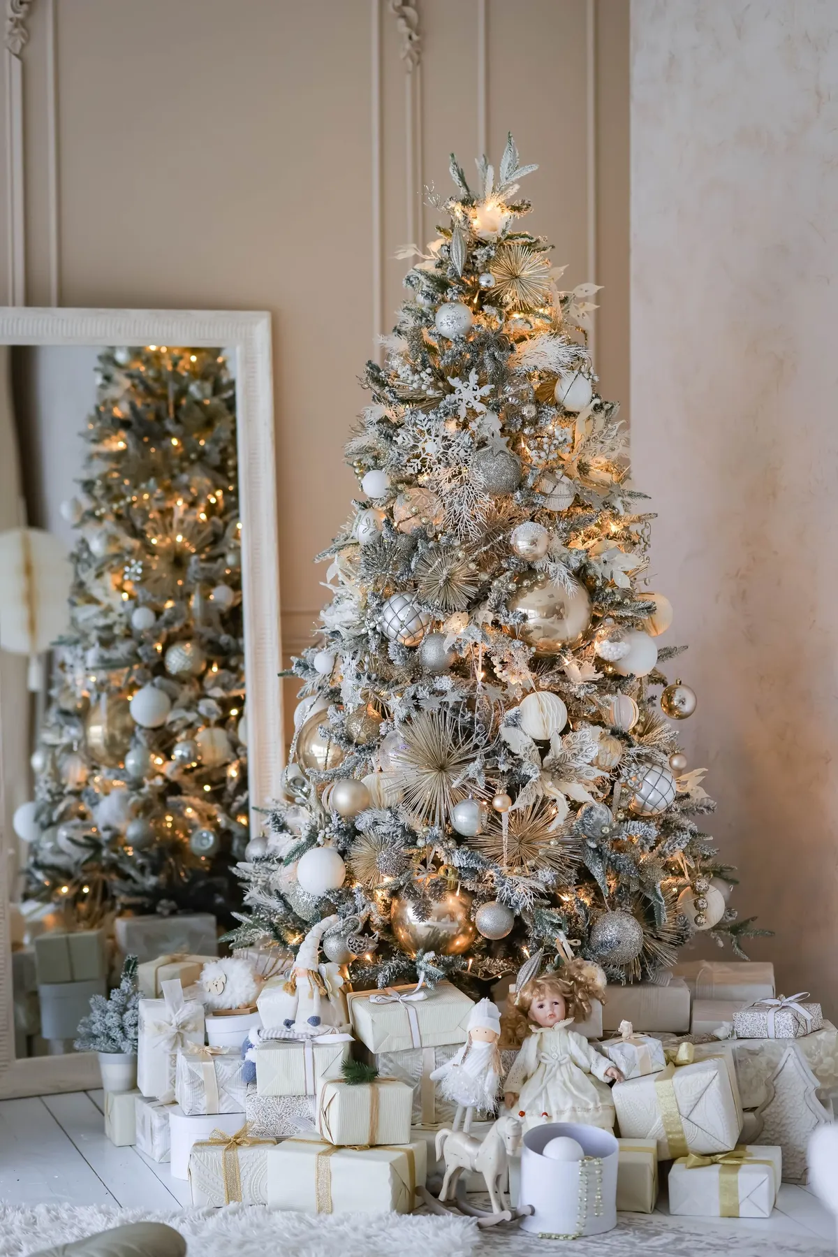 White-flocked tree with ivory, silver and champagne ornaments beside a mirror, with gifts and small toys underneath