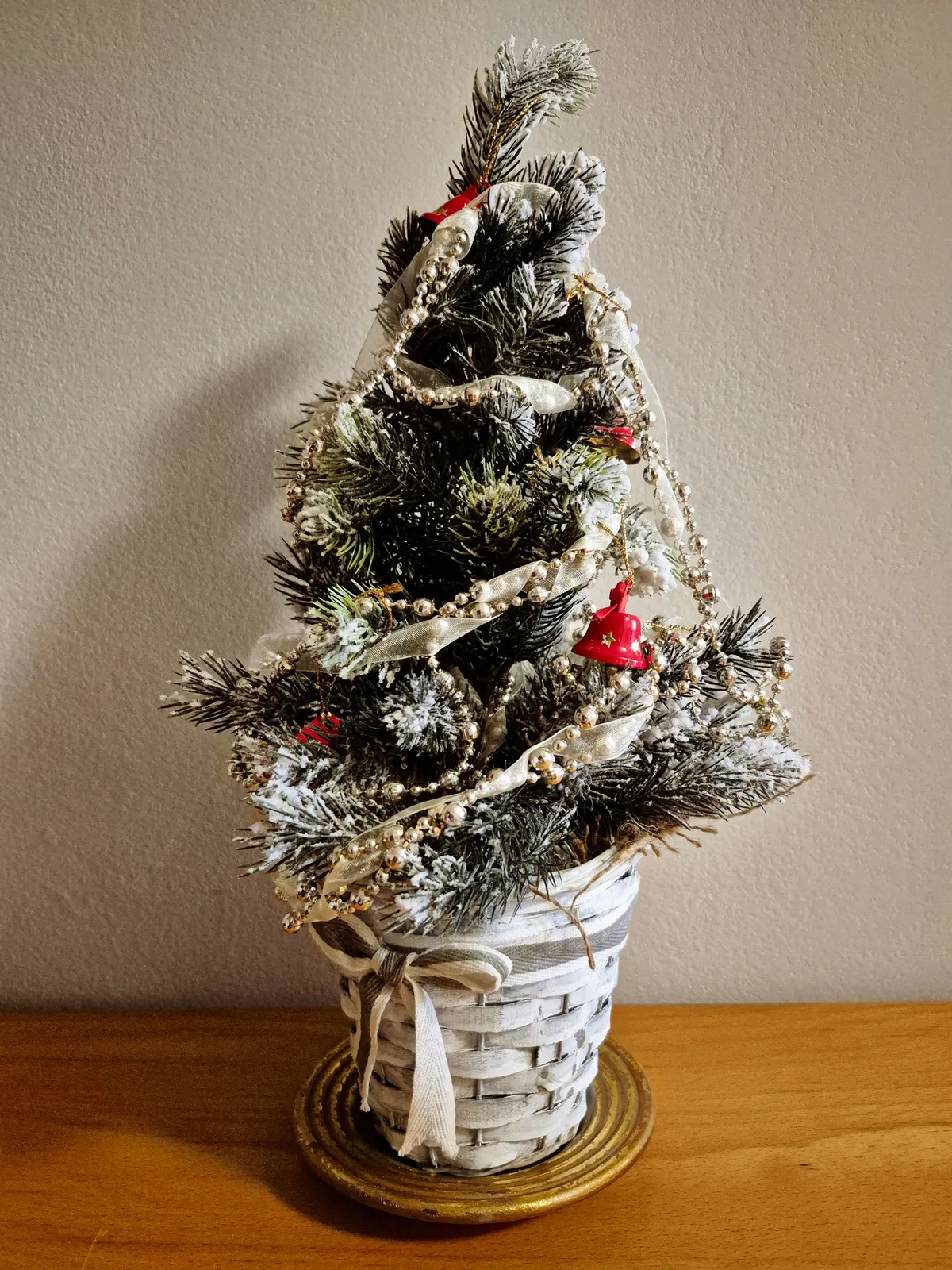 Small flocked tree in a white woven basket with red bells, bead garlands and ivory ribbon spiraling down