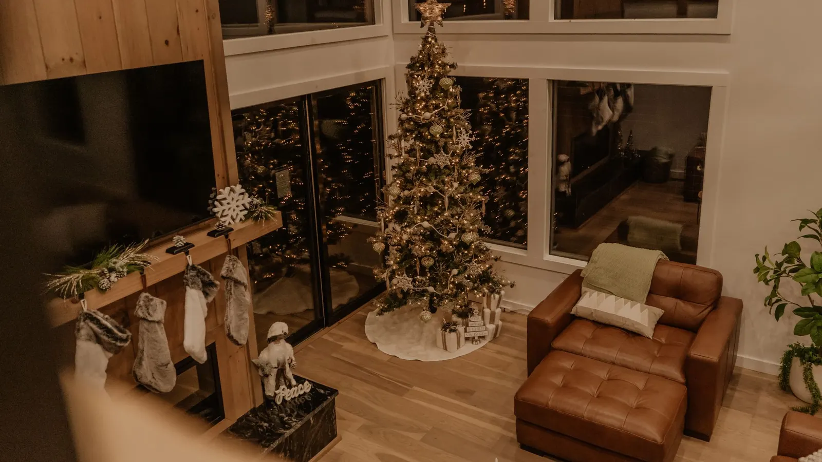 Slim green tree with a gold star, champagne ribbon and white ornaments by tall windows near a mantel with stockings