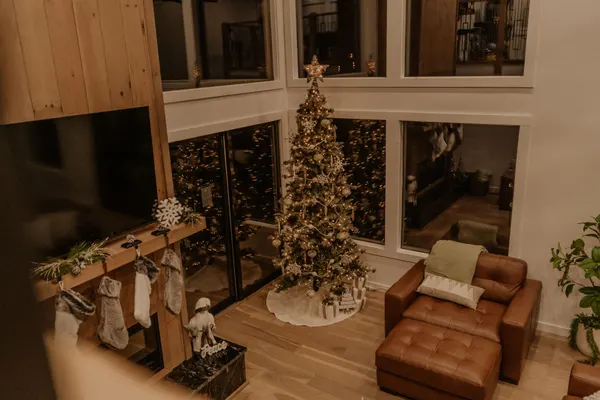 Slim green tree with a gold star, champagne ribbon and white ornaments by tall windows near a mantel with stockings