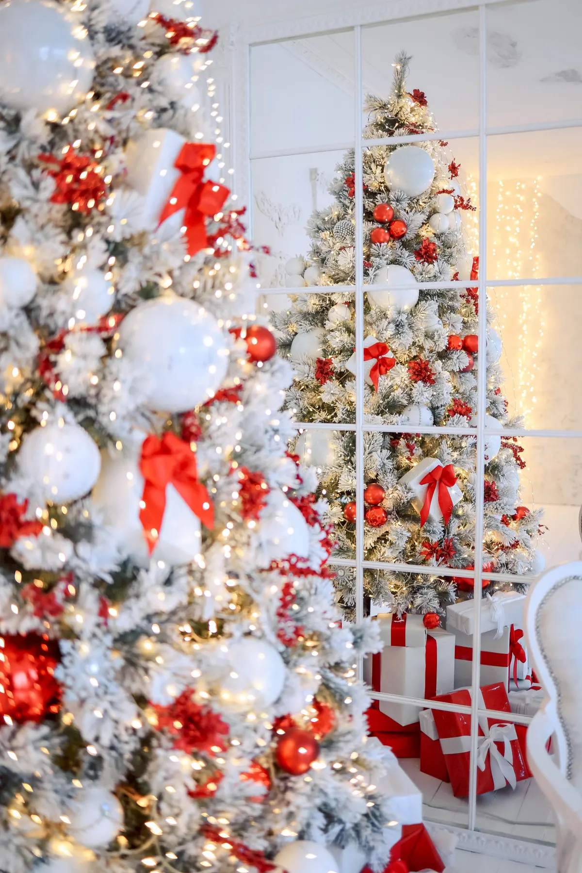 Tall white-flocked tree with red bows, white and silver balls and warm lights reflected in a white mirror