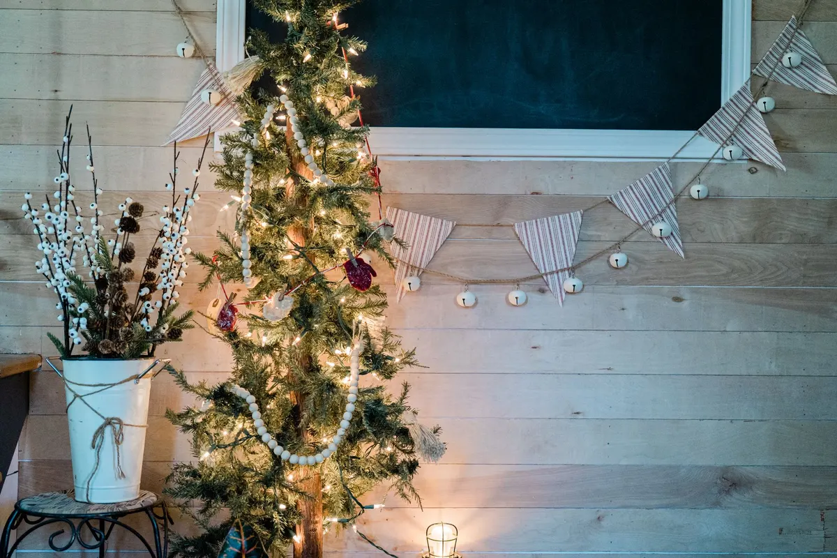 Tall slim sparse green tree with white bead garlands, red mitten ornaments and warm lights beside a twine-tied bucket