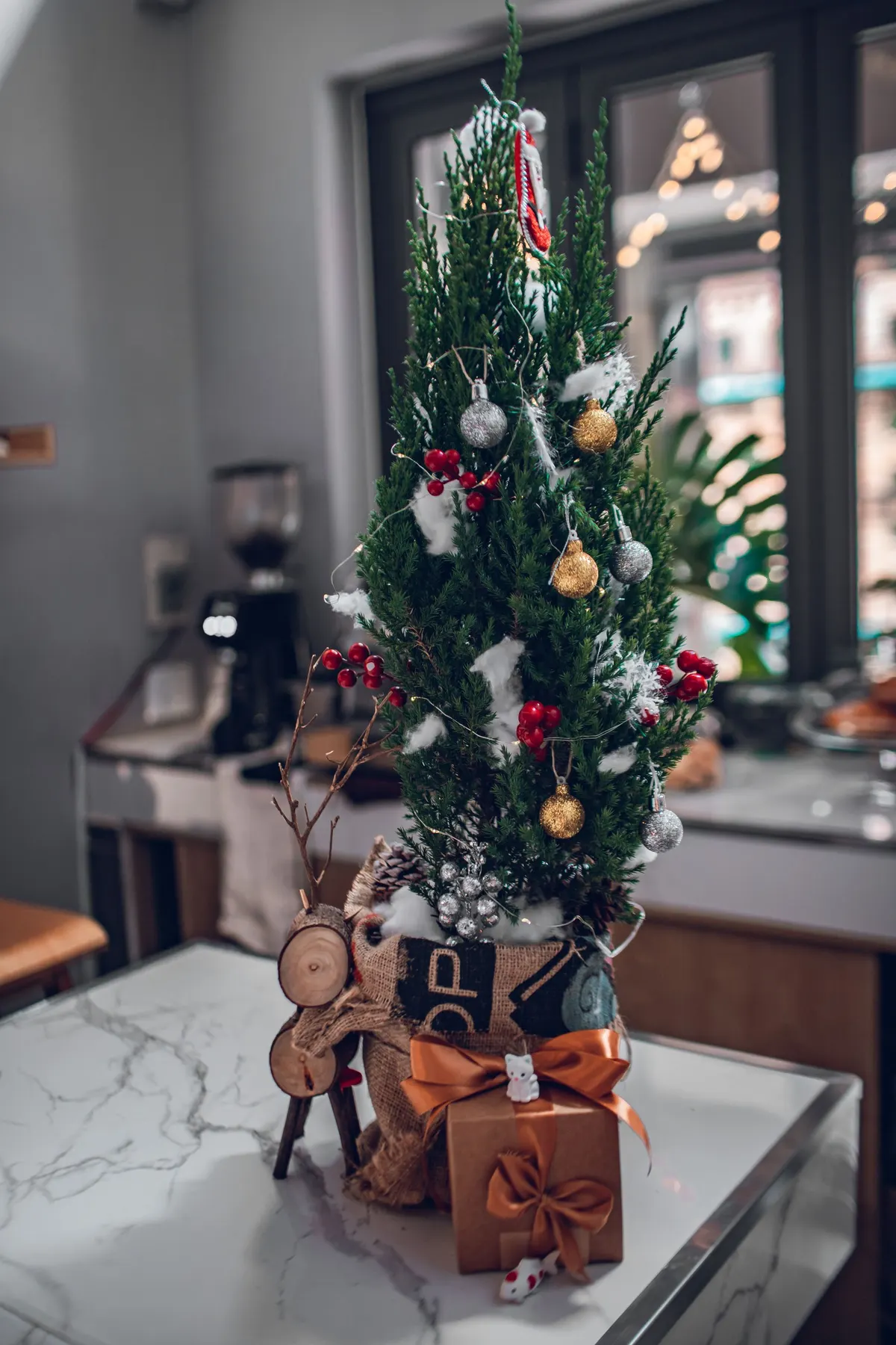 Slim tabletop tree with faux-snow tufts, gold and silver balls, red berry sprigs and copper bows on a marble table