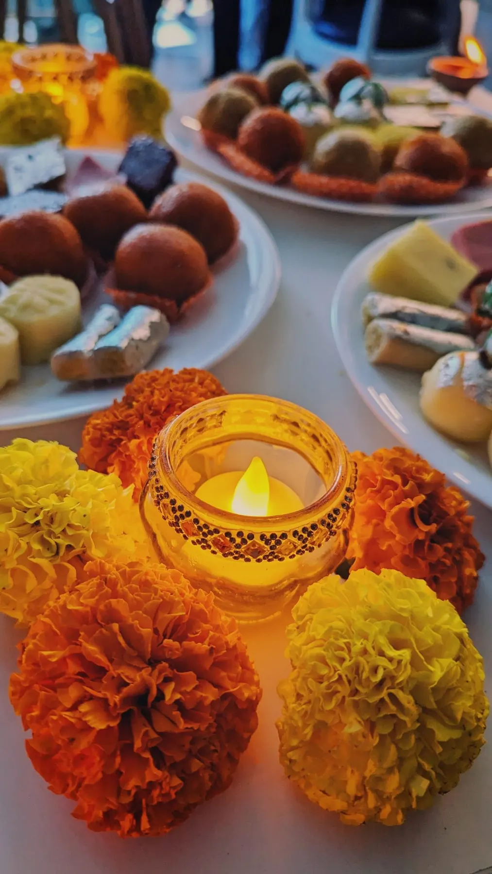 Plates of Indian sweets beside a tealight in a glass holder and orange and yellow marigolds