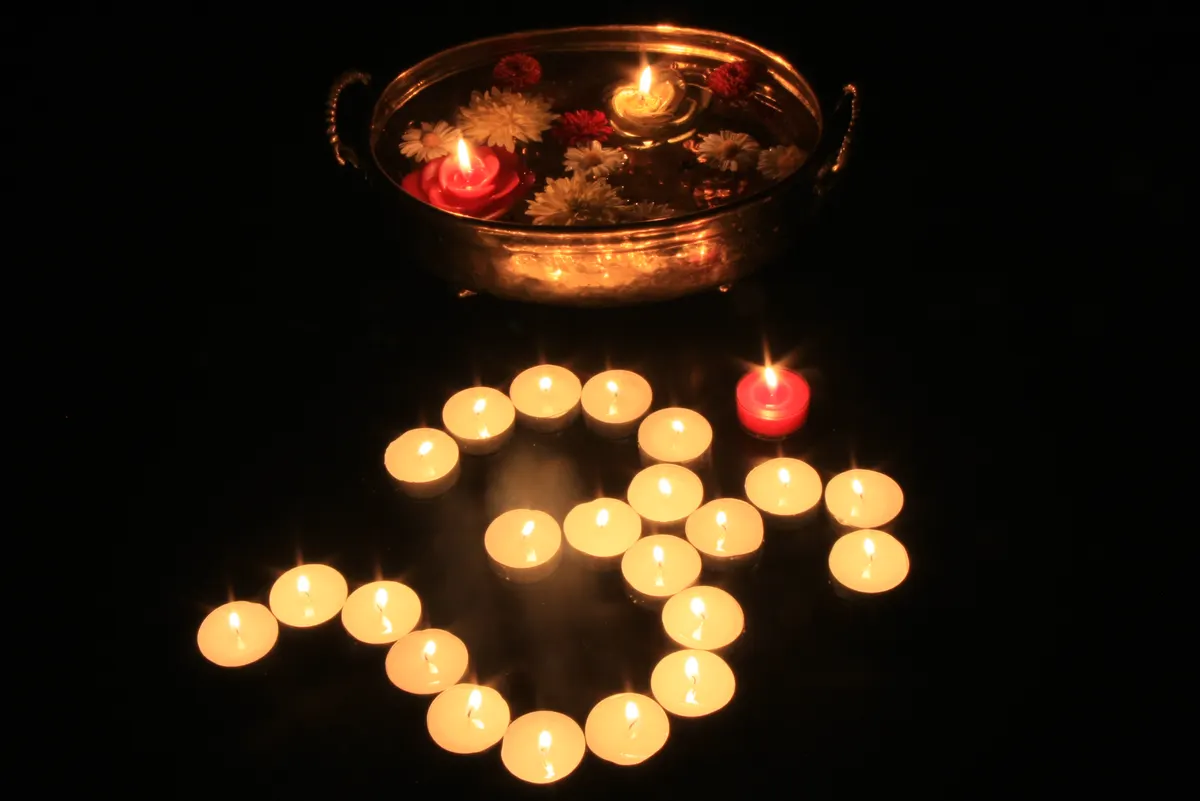Tea-light candles arranged in an Om shape on a dark surface beside a brass bowl of floating flowers and candles