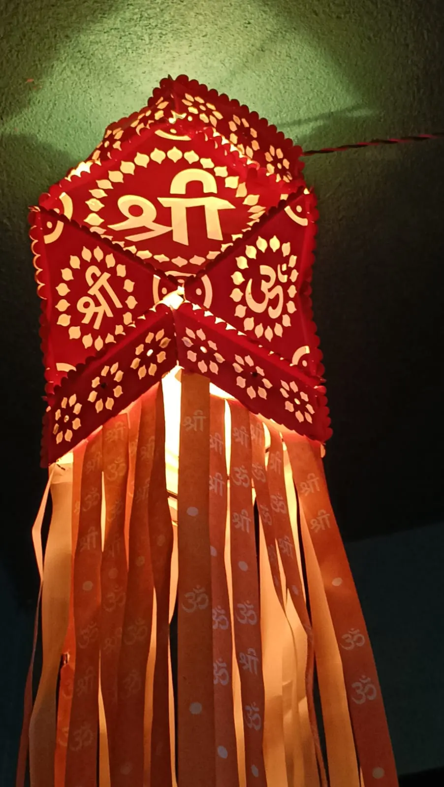 Red paper kandil lantern with a paper fringe, lit from inside against a green wall