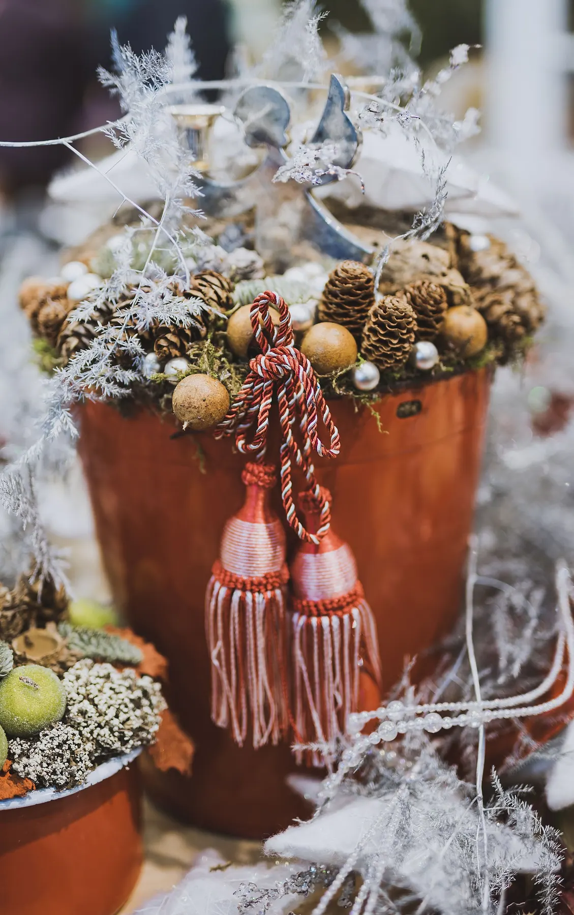 Terracotta pot filled with frosted branches, pinecones, green and gold ornaments and two red knitted tassels, a smaller pot below