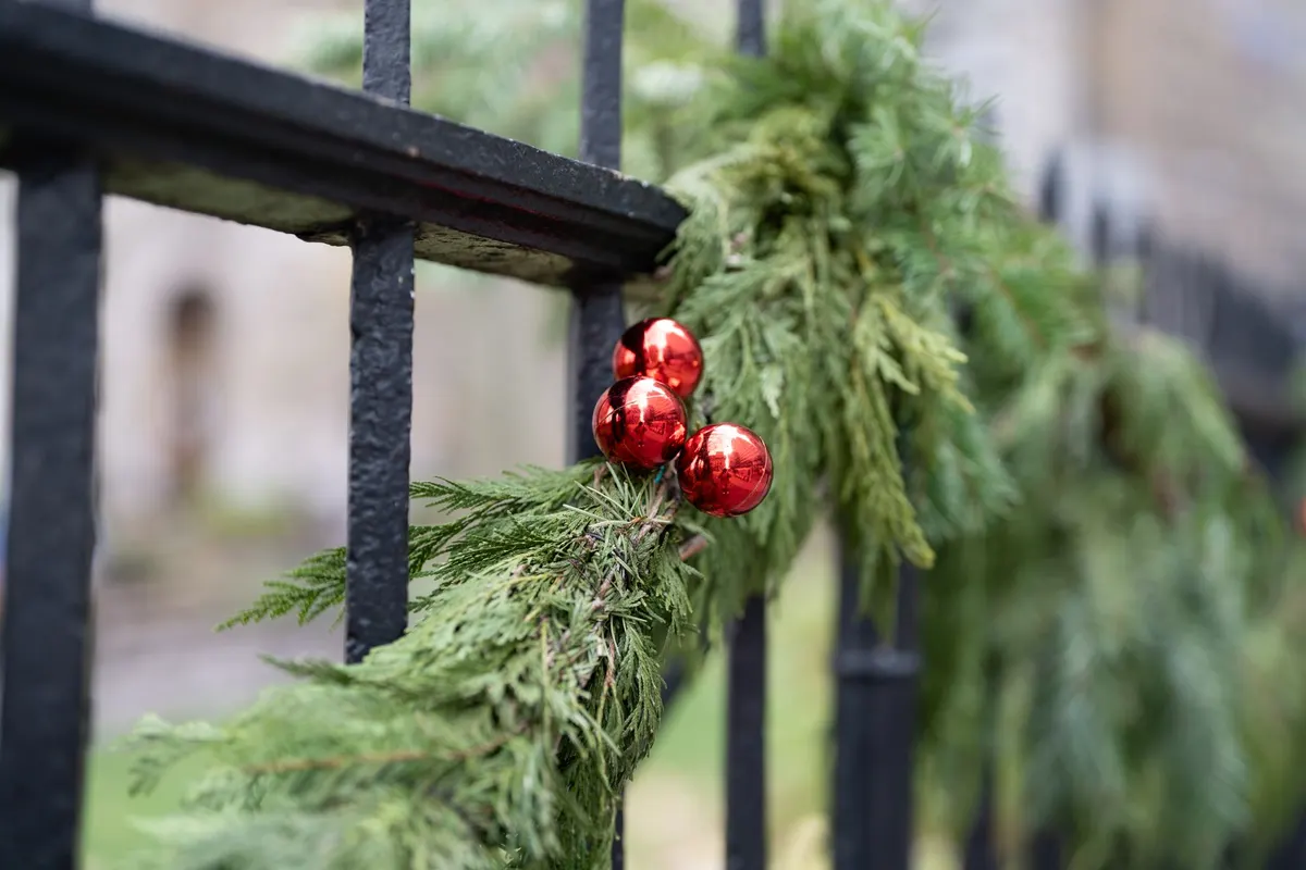 Black iron fence wrapped in evergreen garland with three shiny red baubles, beige buildings blurred behind