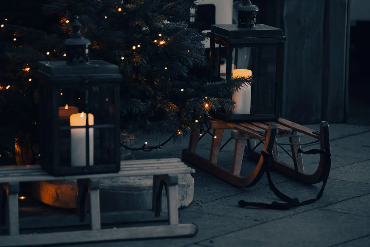 Two black lanterns with cream candles and two wooden sleds among evergreen branches and warm lights on gray stone paving