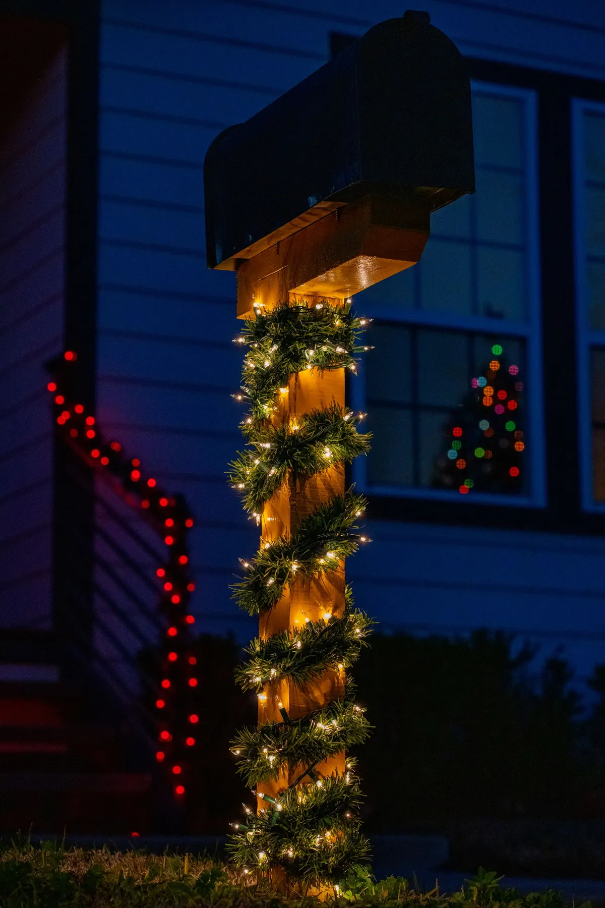 Black mailbox on a wooden post wrapped in green garland and warm lights, with a blue house behind at night