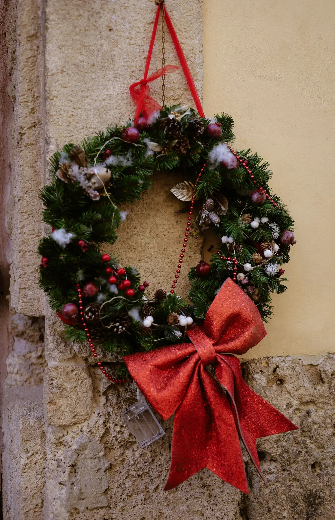 Green wreath with pinecones, red berries, white accents and a large red bow on a beige stone and plaster wall