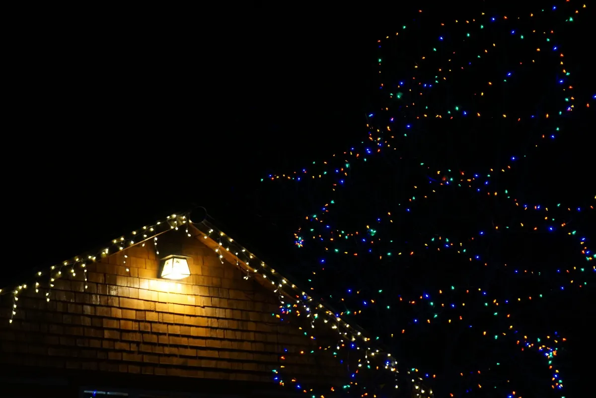 Brown shingled house at night with warm white roofline lights, a yellow wall lamp and a tree in colored lights