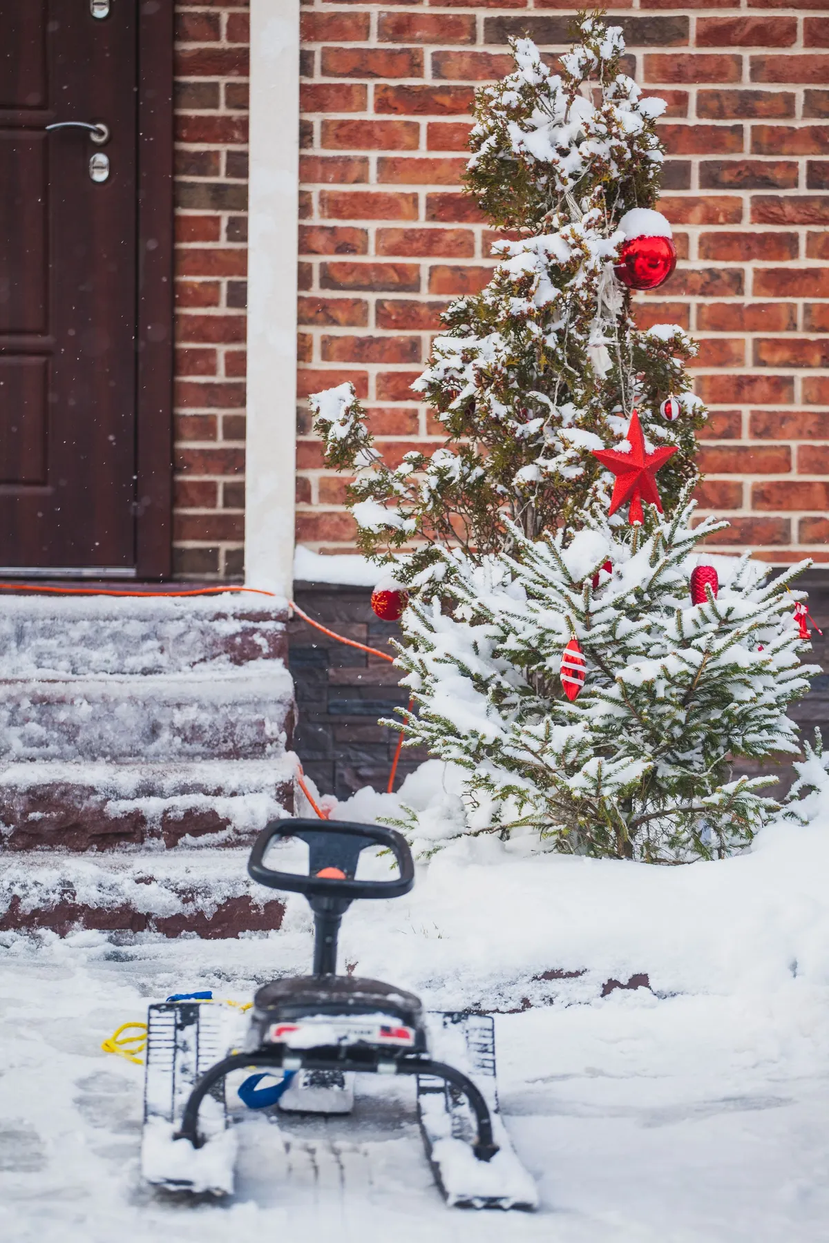 Two small snowy evergreens with red ornaments beside a brown door and snowy steps, with a black sled leaning nearby