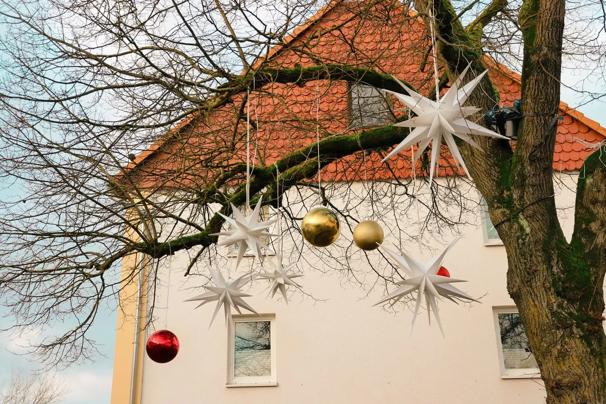 Five white stars, two gold ornaments and a red bauble hanging from bare branches beside a cream house