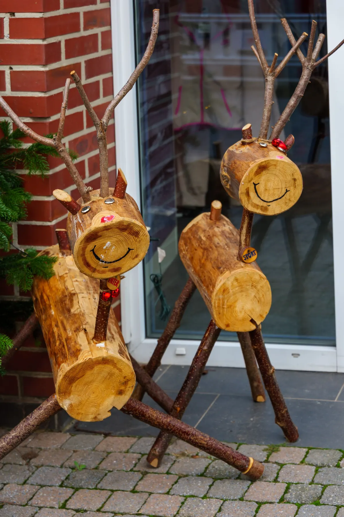 Two wooden reindeer with branch antlers and log bodies stand on gray paving beside a brick wall and glass door