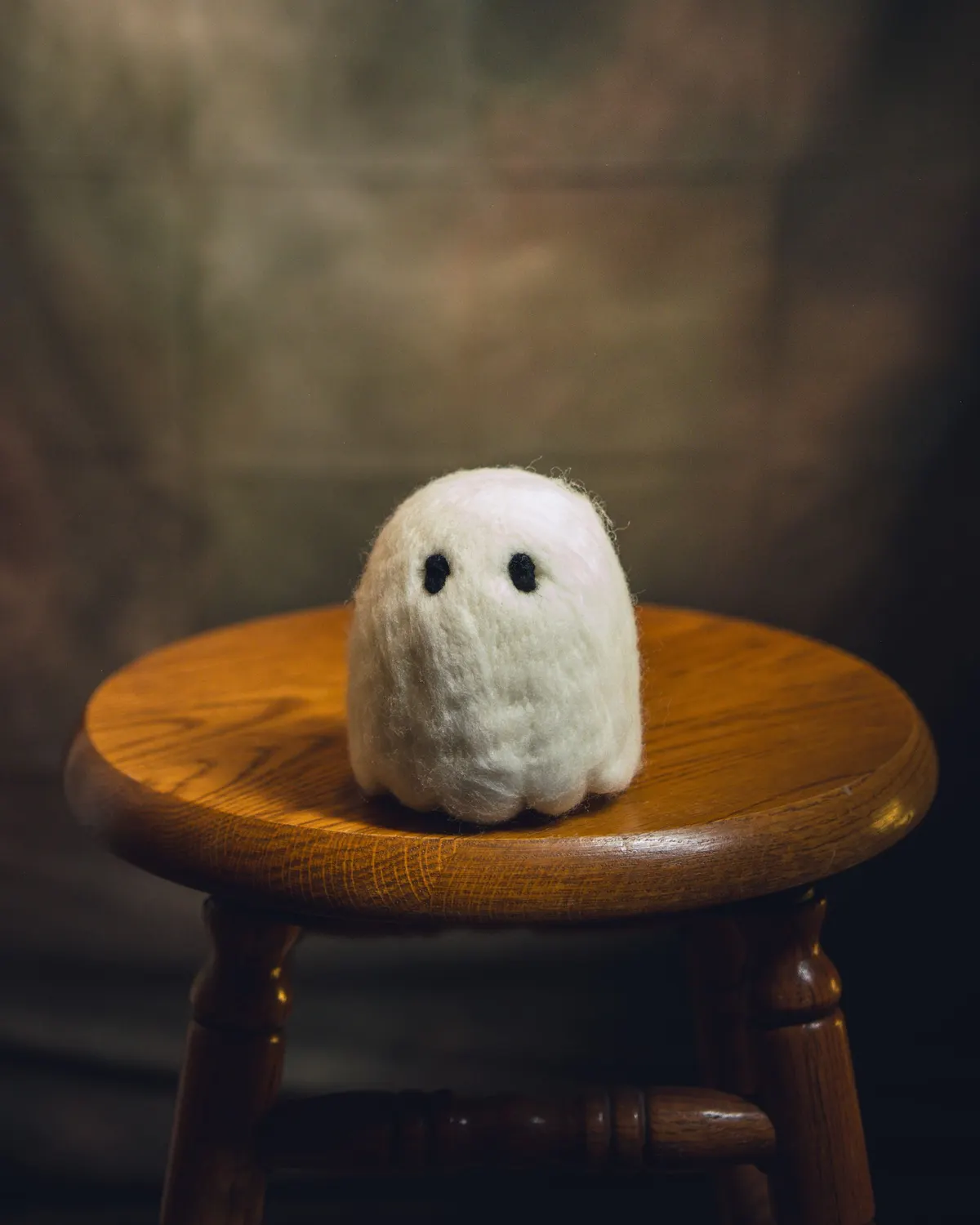Palm-sized needle-felted white wool ghost with two black oval eyes and a scalloped hem on an oak stool