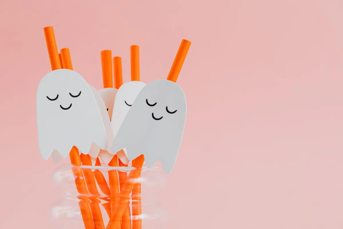 White paper ghosts with sleepy closed eyes on orange paper straws in a rippled clear glass vase