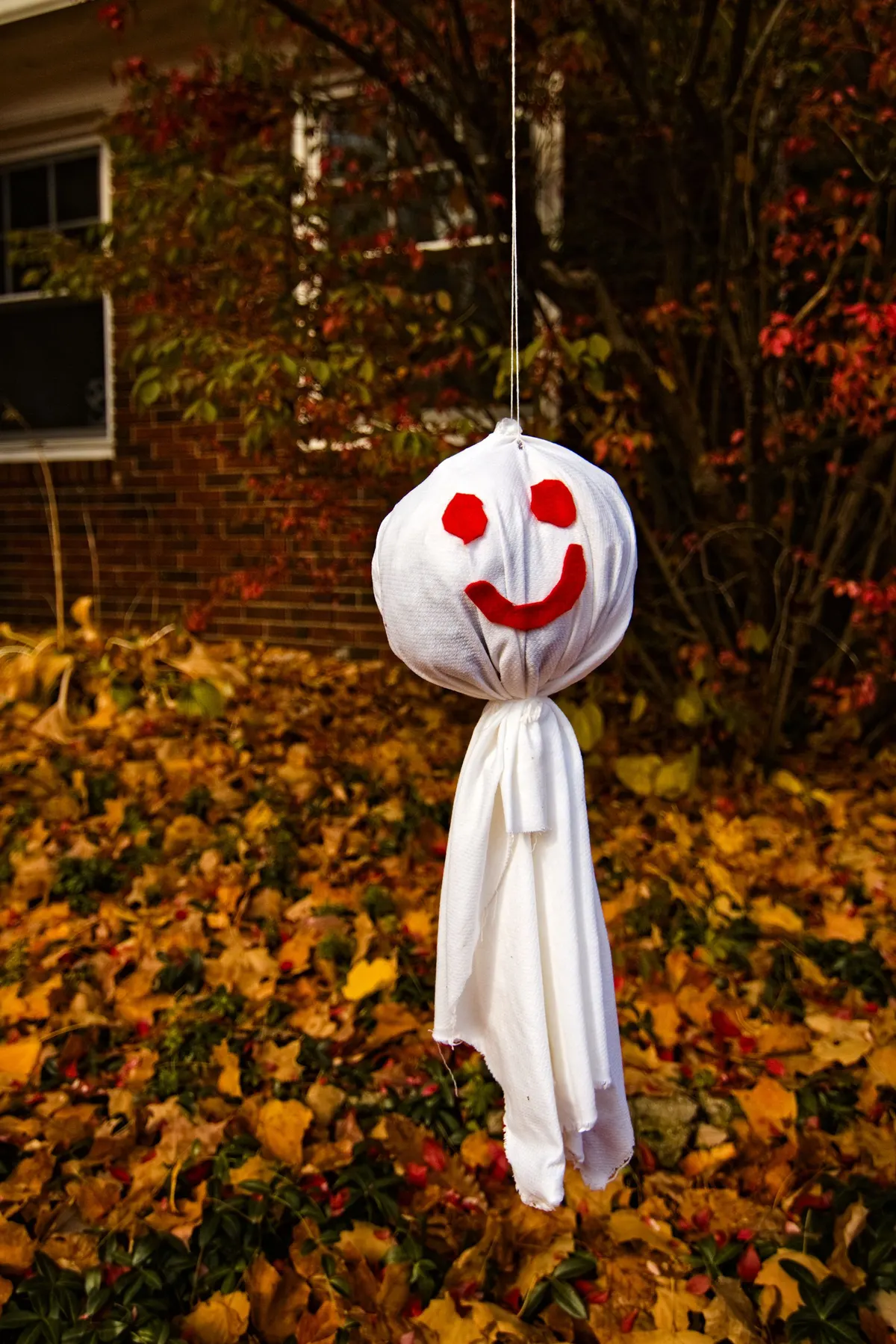 White cloth ghost tied around a ball head with red felt eyes and smile, hanging on a string