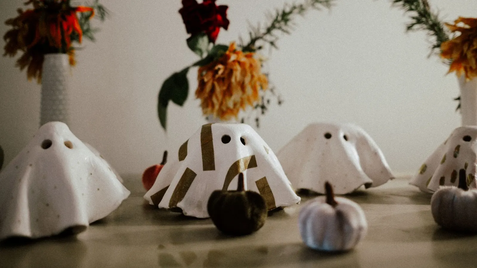 Small stiffened white cloth ghosts with round eye holes, one taped in gold stripes, beside velvet pumpkins and dried flowers
