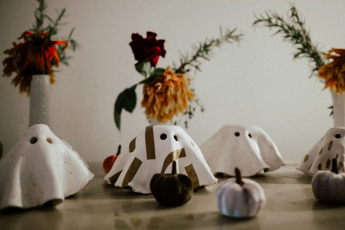 Small stiffened white cloth ghosts with round eye holes, one taped in gold stripes, beside velvet pumpkins and dried flowers