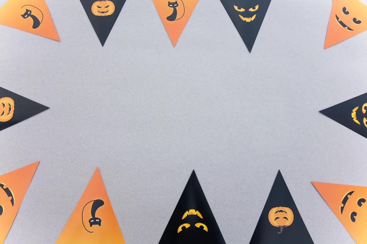 Orange and black paper pennants with black cats, jack-o'-lantern faces and pumpkins laid around a gray surface