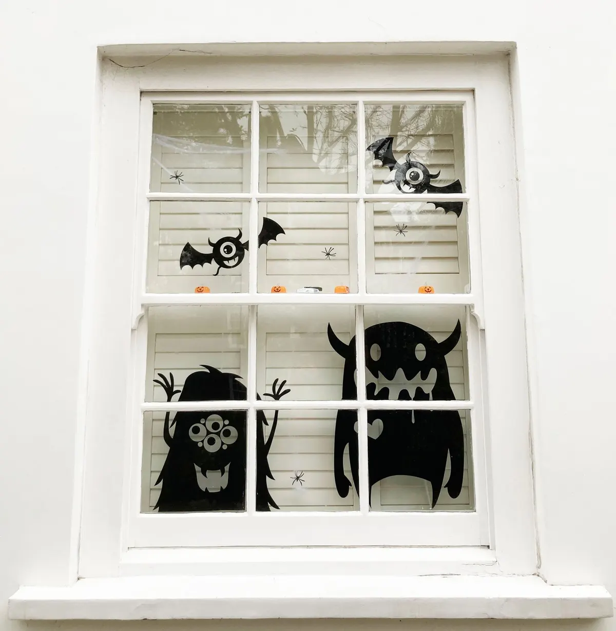 White sash window with black monster, one-eyed bat and spider silhouettes on the glass and tiny pumpkins on the rail